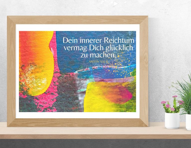 Gerahmtes Bild mit bunter, abstrakter Acrylmalerei und dem Zitat 'Dein innerer Reichtum vermag Dich glücklich zu machen.' von Anton Saur auf einem Regal vor einer grauen Wand.